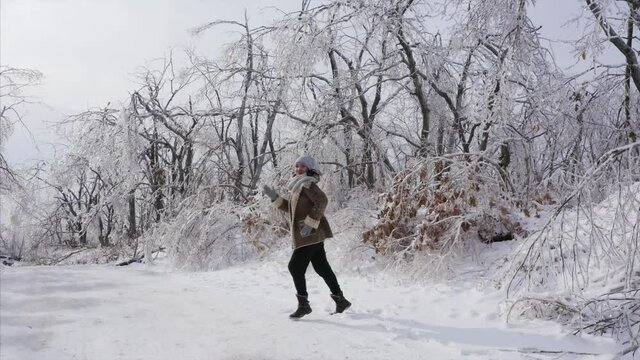 A cheerful young woman dancing on the path in the forest that looks magical because branches are covered with ice after icy rain, natural disaster