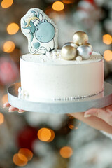 A woman holds a cake in her hands with Christmas decor on the background of a Christmas tree with lights. High quality photo.
