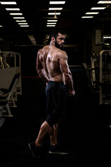 Fototapeta na wymiar strong young bearded caucasian man with sport physique body standing in dark fitness gym showing powerful back muscle