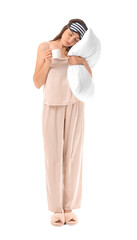 Portrait of young woman in pajamas with sleep mask, pillow and cup of coffee on white background