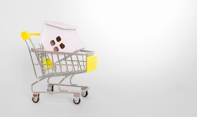 A white wooden house lies in a grocery cart with a yellow handle on a gray background. Side view. Horizontal format. Copyspace