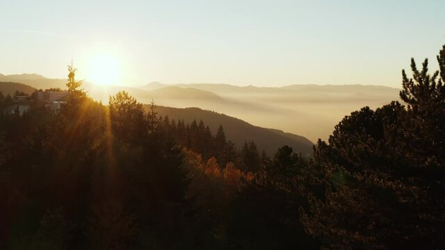 
Drone video of sunset, sunrise in the mountains where you can see mountain ranges with trees in the foreground, the slopes are covered with haze and the sun with rays. Aerial landscape view.