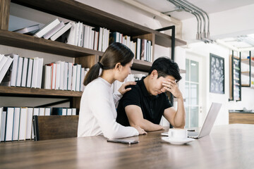 The sad unhappy handsome man sitting in front of a laptop, holding his forehead while having a headache with a woman cheering up by touching his shoulder.