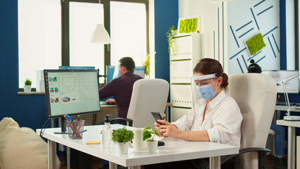 Woman entrepreneur with protective mask working in professional workspace typing on mobile phone in front of computer. Multiethnic coworkers working respecting social distance in financial company