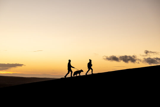 Silhouette of Man and Woman couple walking their dog at sunset on a hill with beautiful clouds and orange hue