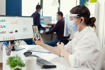 Entrepreneur talking with businesspeople during video call using smartphone at workplace wearing face mask against covid19. Multiethnic coworkers working respecting social distance in business during