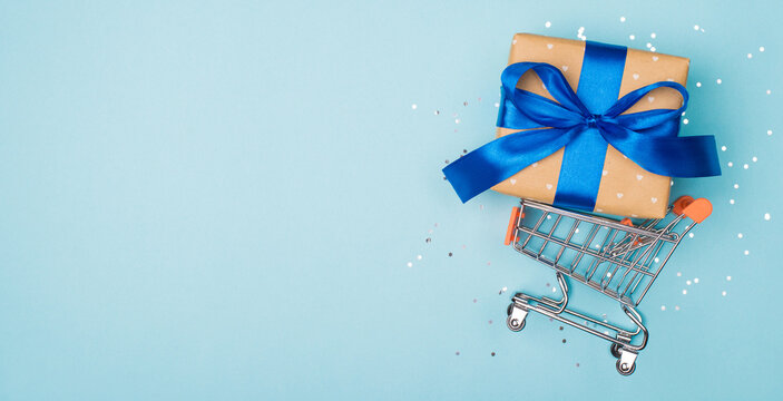 Wide horizontal photo flatlay image of small mini metal pushcart riding and holding large giftbox isolated on light pastel color background with shine glowing shimmer with blank empty space