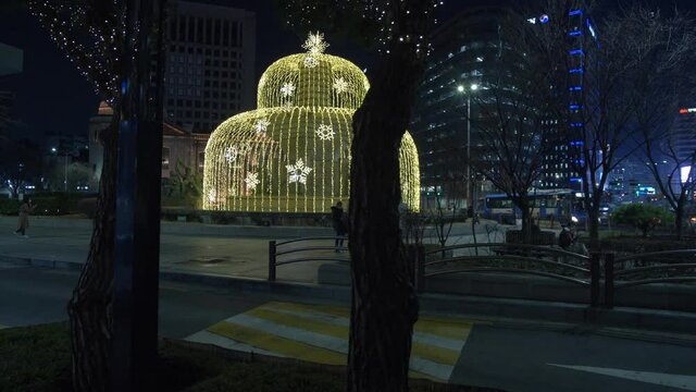 A Christmas light in the shape of a fountain in shopping district of Seoul. Silhouetted trees and road in FG. 4K 60fps gimbal moves along sidewalk from right to left. 