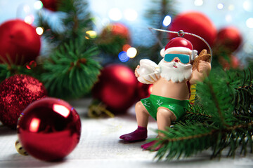 Christmas toy in the shape of Santa Claus in a bathing suit and flippers stands in Christmas tree branches against a background of red Christmas balls and lights on a white board