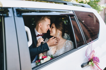A beautiful bride with a bouquet of flowers in a lace dress and a groom in a blue suit are sitting and hugging inside a white car. Wedding portrait of the newlyweds in the window.