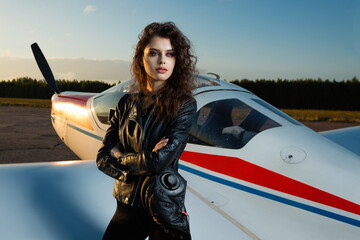 Beautiful sexy girl in a leather flight suit on a sunny day with a sunset sky on the background of...
