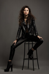 Beautiful sexy girl in black leather pants and a leather jacket posing while sitting on a high bar stool in the studio on a gray background