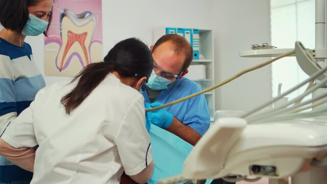 Stomatologist with face mask and gloves using drill tool for cleaning teeth of little patient working together with man assistent in modern stomatological clinc. Mother visiting dentist with child.