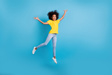 Fototapeta na wymiar Full length photo of little cute afro american girl jump air raise hands wear jeans yellow t-shirt isolated on blue color background