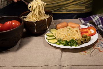 A man sitting at a table from a bowl puts spaghetti into a plate. Sausages, peppers, cucumbers and olives are on the plate.