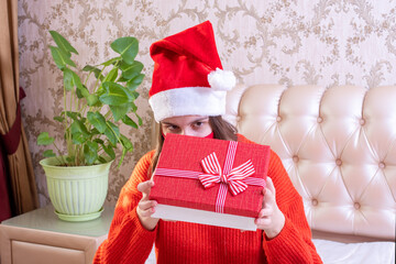 Young happy caucasian girl wearing santa claus hat and red protective mask opens a gift box for celebration, merry christmas and happy new year. Christmas stay at home concept
