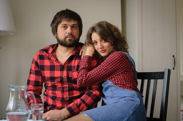 Lovely couple wearing similar red checkered shirts is hugging sitting at the table at home during celebration St. Valentine Day. Indoor portrait of boyfriend and girlfriend