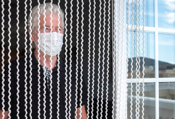 Coronavirus. Close-up of a senior man white-haired at home behind a curtain in self isolation wearing a protective face mask due to coronavirus,  looking the world from window