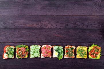 Various sandwiches on toast bread with salmon, sprouts, vegetables, egg lie in a row on a brown wooden background. Healthy food concept.