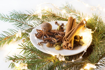 spices for mulled wine on a small white plate on a white background with green branches of a Christmas tree and a burning garland of warm yellow color.