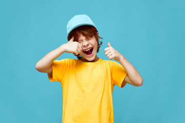 Fototapeta premium Cheerful red-haired boy with open mouth holds thumbs up yellow t-shirt cap 