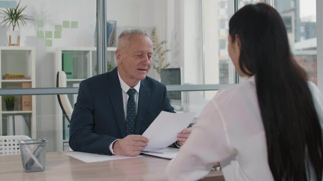 Medium close-up over shoulder of aged Caucasian businessman sitting by desk, smiling, talking to unrecognizable long-haired candidate passing him paper with CV on job interview