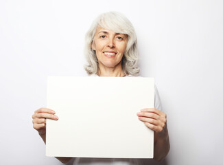 Fototapeta premium Elderly grey-haired woman with blank advertising board or copy space. Portrait of handsome expressive grandmother over white background.