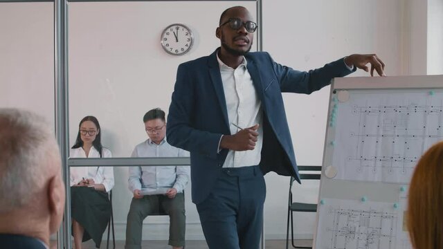 Medium Long Of Young African Businessman In Blue Suit Standing By Whiteboard With Graphs On It, Making Presentation For Colleagues In Office, Two People Waiting Outside Room Behind Glass Wall