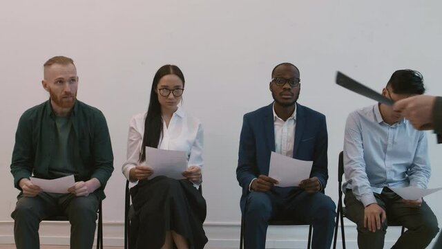 Medium long shot of four diverse impatient people sitting on chairs in hallway, holding resumes in hands, unrecognizable manager inviting young Asian female candidate for interview
