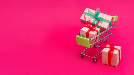 Small grocery cart with gift boxes on red-pink background. Gifts for Valentine's day, Christmas and birthday. Shopping online. Holiday sales and discounts for New year. Retail and wholesale purchases