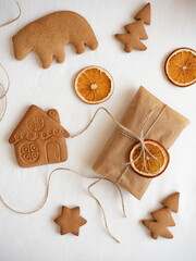 Gingerbread cookies of different shapes, gift wrapped in craft packaging, dried orange slices laid out on a white tablecloth in the morning light, top view