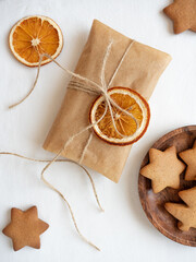Gingerbread cookies of different shapes, gift wrapped in craft packaging, dried orange slices laid out on a white tablecloth in the morning light, top view