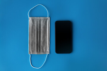 Top view mockup grey surgical mask for prevent from coronavirus COVID-19 pandemic next to blank screen mobile phone on plain blue isolated background. Healthcare and technology concept