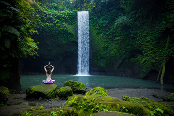 Fototapeta na wymiar Young Caucasian woman meditating, practicing yoga at waterfall. Lotus pose. Hands raised up in namaste mudra. Tibumana waterfall, Bali, Indonesia. View from back.