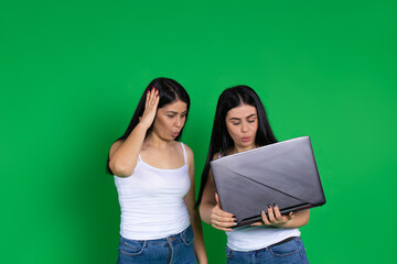 Surprised twin women in casual clothes look at laptop. Photo on green background with side space.