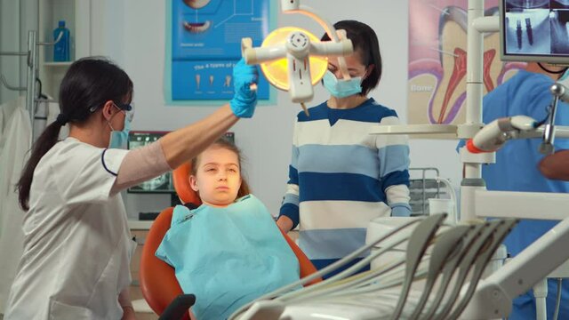 Pedicatric dentist doctor working in dental unit with nurse and little girl patient. Stomatologist speaking to mother of girl with toothache sitting on stomatological chair while man preparing tools.