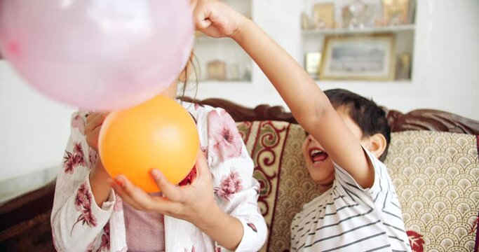 Slow-motion close of a boy and a girl playful fun enjoy naughty love care share inflating color balloons in living room on couch and fight hitting each other in between looking at camera pov