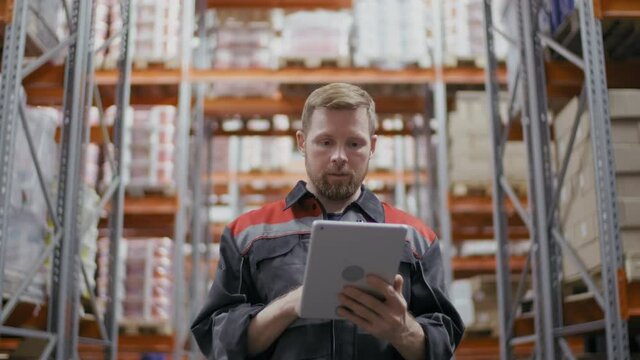 Low-angle medium footage of caucasian construction supervisor in blue coverall using digital tablet while controlling storage goods looking at shelves at big warehouse