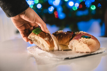 a hand picking up a piece of roscón de reyes, ring pastry roll, with an illuminated Christmas tree. Christmas concept and typical food of Spain
