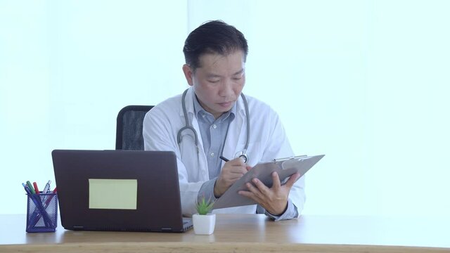 Doctor Filling Medical History Sheet And Using Laptop At Workplace