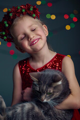 Christmas portrait happy young girl holding cat. Wearing red sequin dress and Christmas wreath. Blond hair and blue eyes posing and fool around in studio on grey background with boke lights.