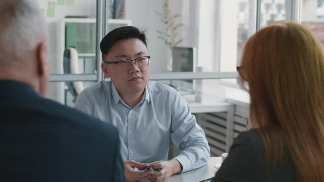 High angle chest-up of smart Asian male candidate sitting by desk in office in front of two unrecognizable company workers on job interview, nodding, listening