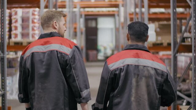 Back-view medium footage of couple of workmen in blue overalls working together chatting while walking along big warehouse