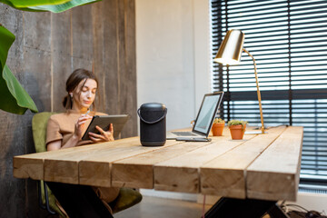 Woman working on the digital tablet at cozy home office. Smart speaker on the table. Concept of a smart home and work from home