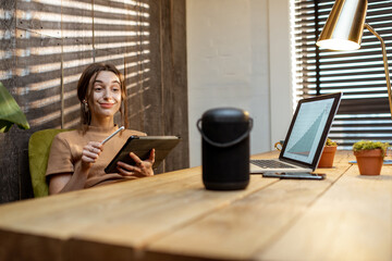 Woman talking to the smart speaker while working on the digital tablet at cozy home office. Concept...
