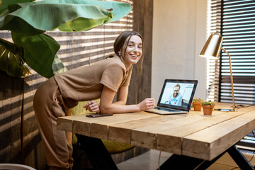 Portrait of a young and cheerful woman in domestic suit having a video connection with colleagues, working on laptop from a cozy home office