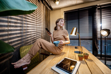 Relaxed woman in comfortable domestic suit with a smart phone and coffee cup sitting on the table at the beautiful home office