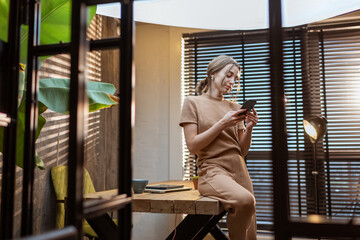 Relaxed woman in comfortable domestic suit with a smart phone and coffee cup sitting on the table at the beautiful home office