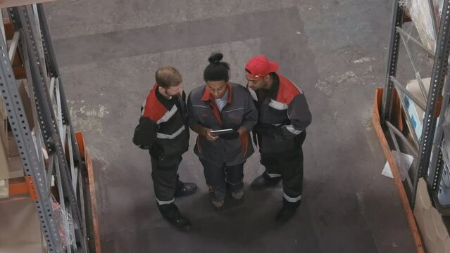 Top-view shot of two multi-ethnic men and one african american woman working together in big stockroom wearing blue coveralls looking at tablet discussing workplan