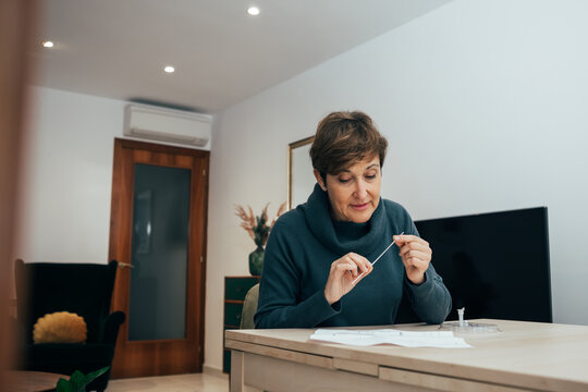 Senior woman sitting at home, studying the instructions for a self test for COVID-19 with Antigen kit on hand. Holds a nasal swab for possible infection of Coronavirus. Health services online.